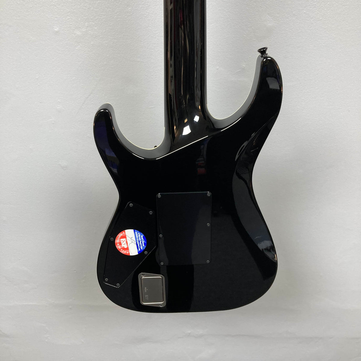 Back view of an ESP E-II Horizon FR-7 Black w/Case 7-string electric guitar by ESP, featuring two rectangular panels and a round inspection sticker on the lower left, hanging against a white wall.