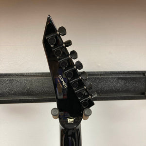 Close-up of the ESP E-II Horizon FR-7 Black electric guitar headstock, showing six tuning pegs and serial number ES176969, resting on a wall-mounted holder. Comes with case. Brand: ESP.