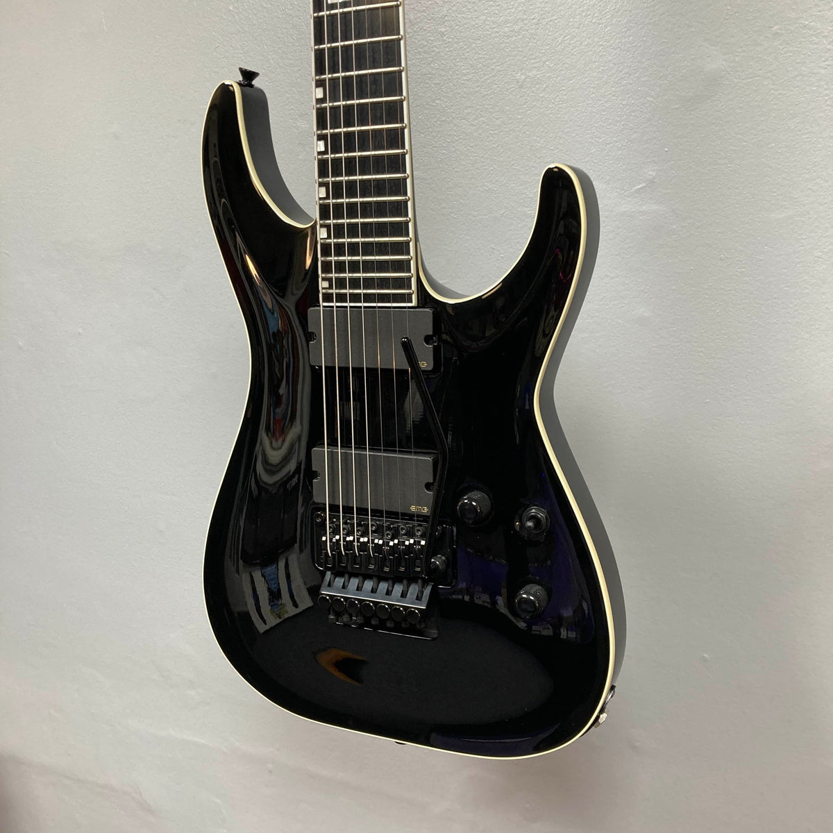An ESP E-II Horizon FR-7 Black 7-string electric guitar with a gloss finish, white binding, humbuckers, Floyd Rose bridge, and three knobs hangs against a white wall. Comes with case.