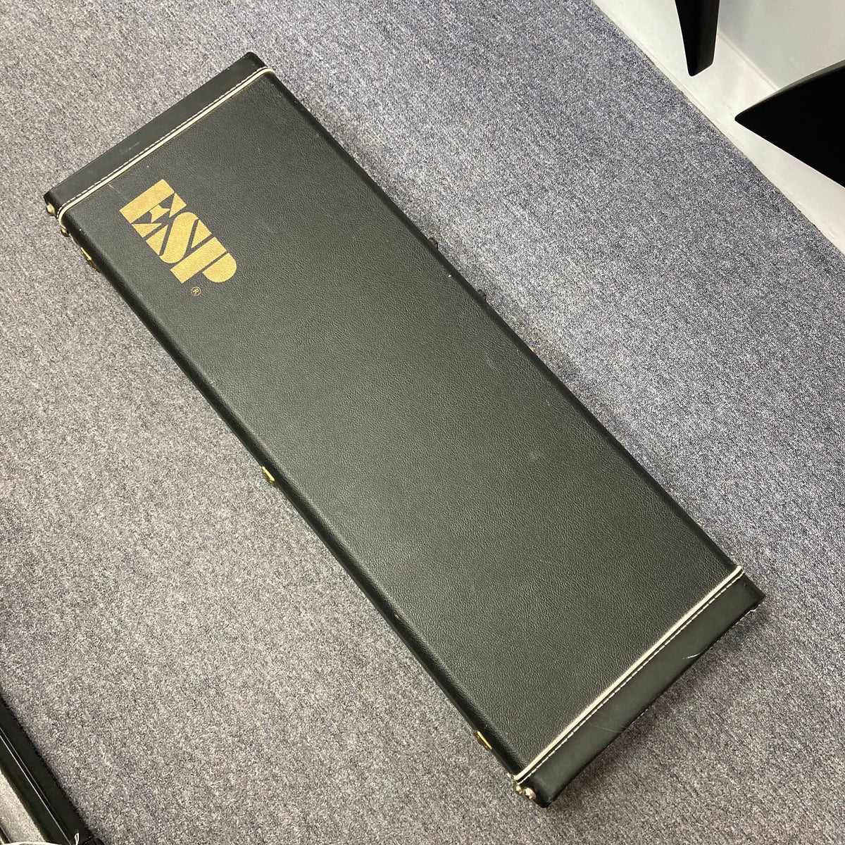 A black rectangular hard case with gold "ESP" lettering, designed for the ESP Standard Eclipse Transparent Cherry Red guitar by Esp, is shown resting on a gray carpeted floor.