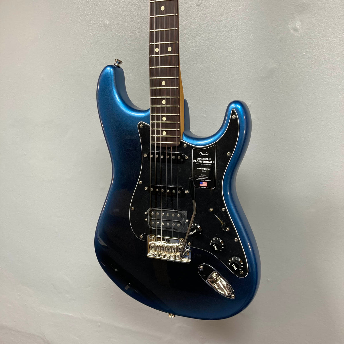 A Fender American Professional II Stratocaster HSS in Dark Night, featuring a black pickguard and three pickups, hangs on a light gray wall.