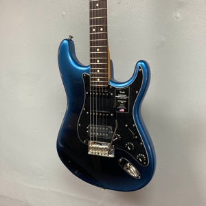 A Fender American Professional II Stratocaster HSS in Dark Night, featuring a black pickguard and three pickups, hangs on a light gray wall.