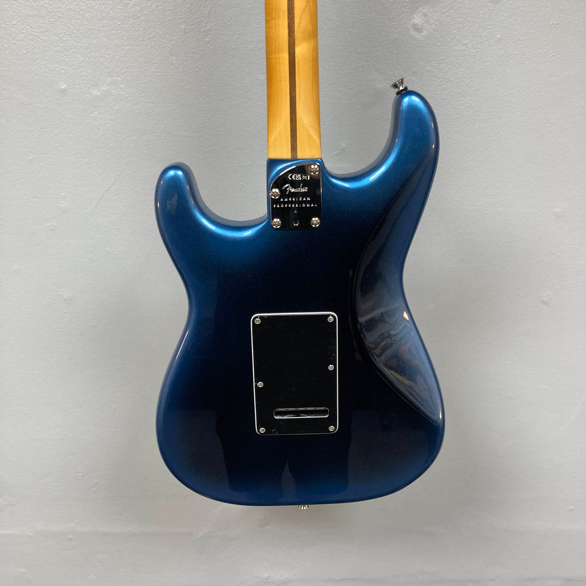 A Fender American Professional II Stratocaster HSS in Dark Night hangs on the wall, shown from the back to display its body, neck, and metal plate with screws.