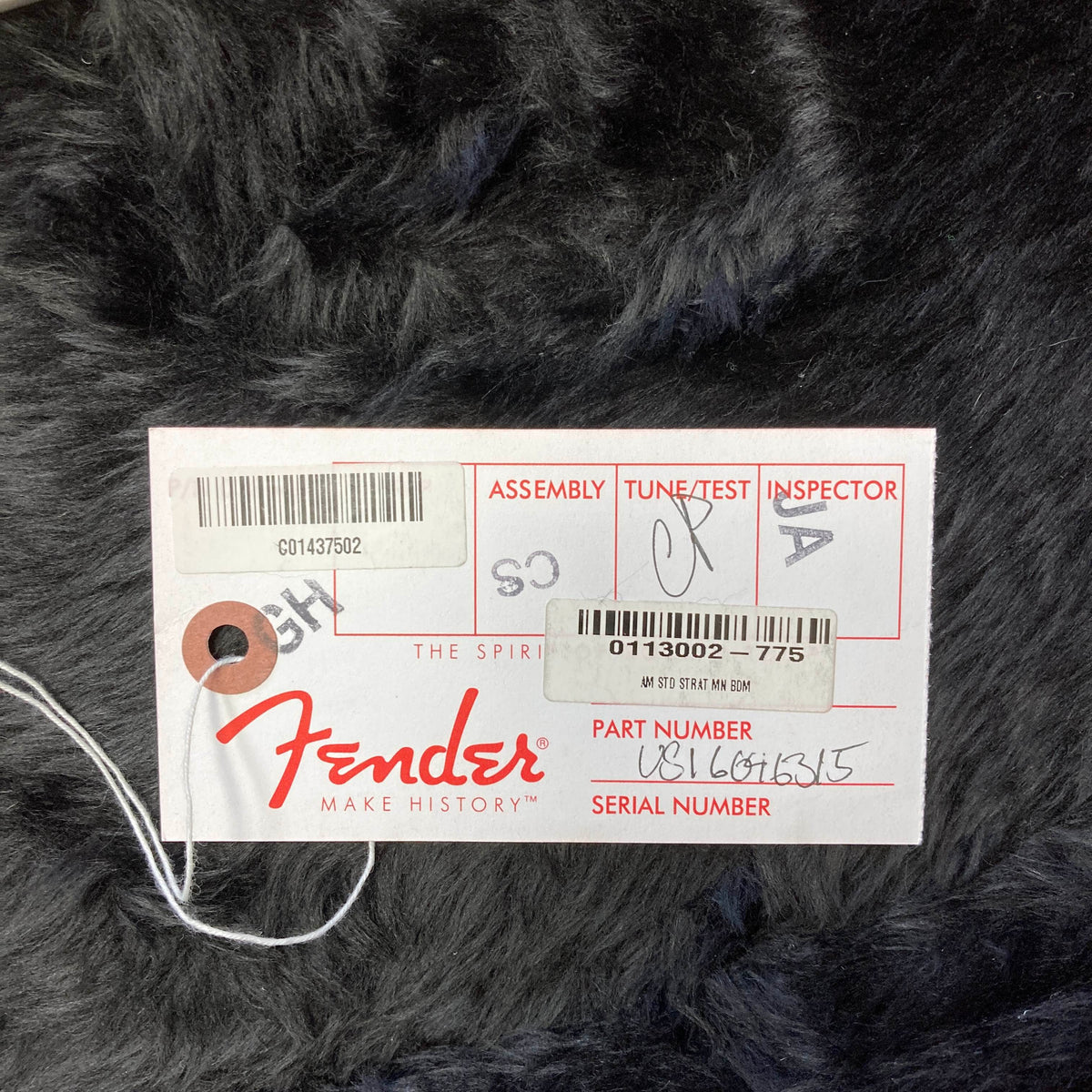 A white Fender inspection tag with handwritten signatures and numbers rests on a black fur-like surface, attached by a string—an authentic detail included with this Fender American Standard Strat upgraded Gilmour EMG DC-20 in the striking Bordeaux Metallic finish (w/Case, Used).