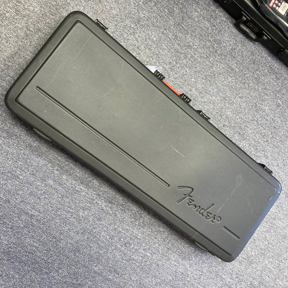Black rectangular hard guitar case with "Fender" logo, lying closed on a gray carpeted floor—perfect for protecting your Fender American Standard Strat upgraded with Gilmour EMG DC-20 pickups in stunning Bordeaux Metallic.
