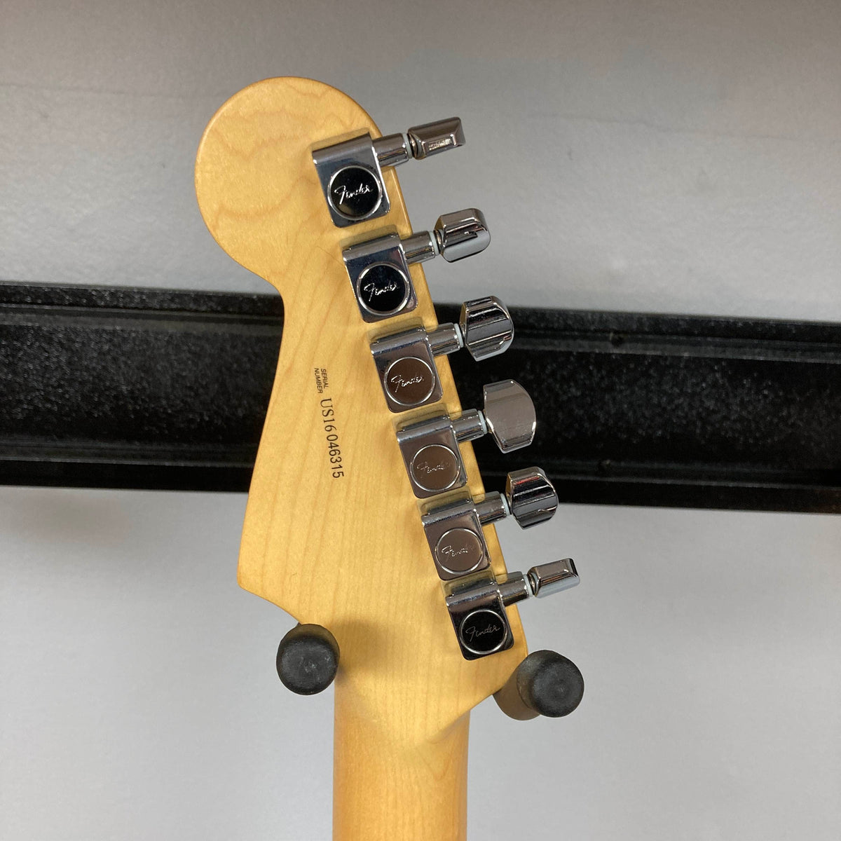 Close-up of the back of a Fender American Standard Strat upgraded Gilmour EMG DC-20 Bordeaux Metallic headstock, showing six chrome Fender tuning pegs and a visible serial number, resting on a black wall-mounted guitar hanger.