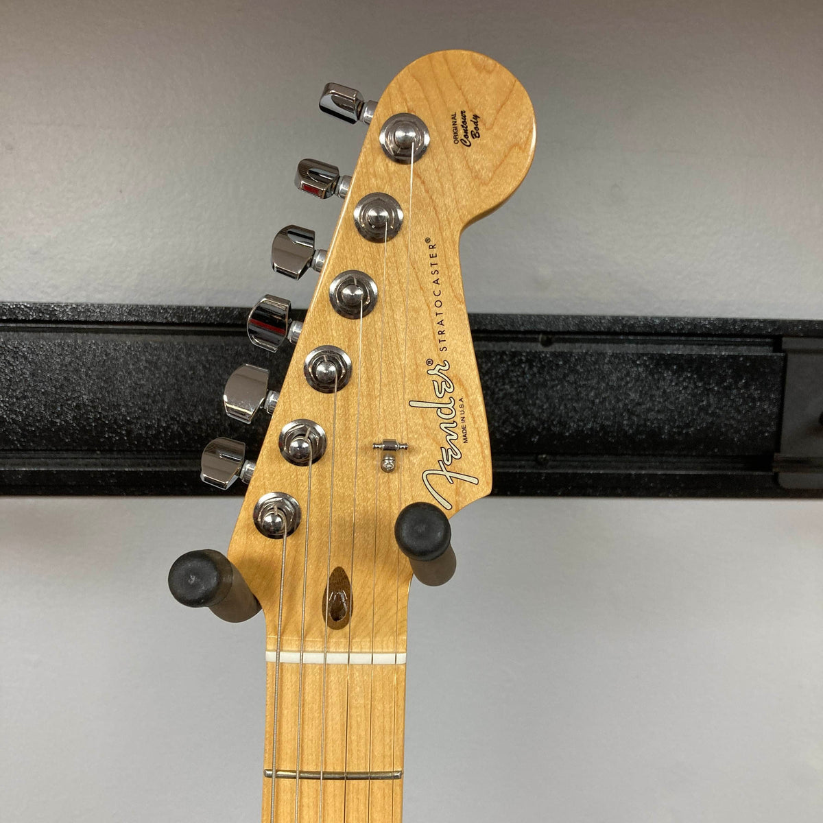 Here’s a rewritten sentence using your product data: Close-up of a Fender American Standard Strat upgraded Gilmour EMG DC-20 in striking Bordeaux Metallic, showcasing six tuning pegs, string guides, and the classic Fender logo. This used guitar comes with a case and is displayed on a wall hanger against a gray background.