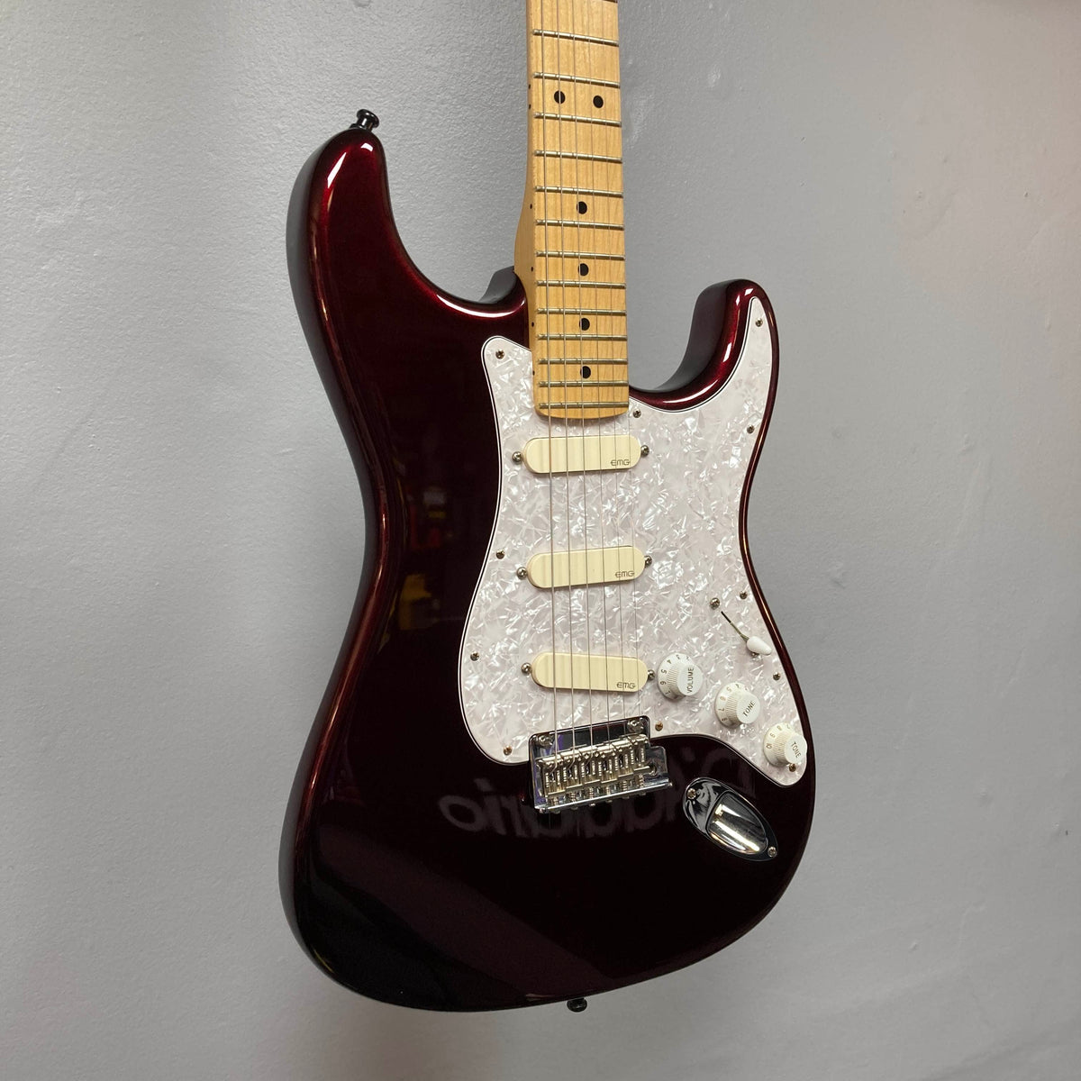 A Fender American Standard Strat upgraded with Gilmour EMG DG-20 pickups in Bordeaux Metallic, featuring a white pickguard, is hanging on a plain gray wall.