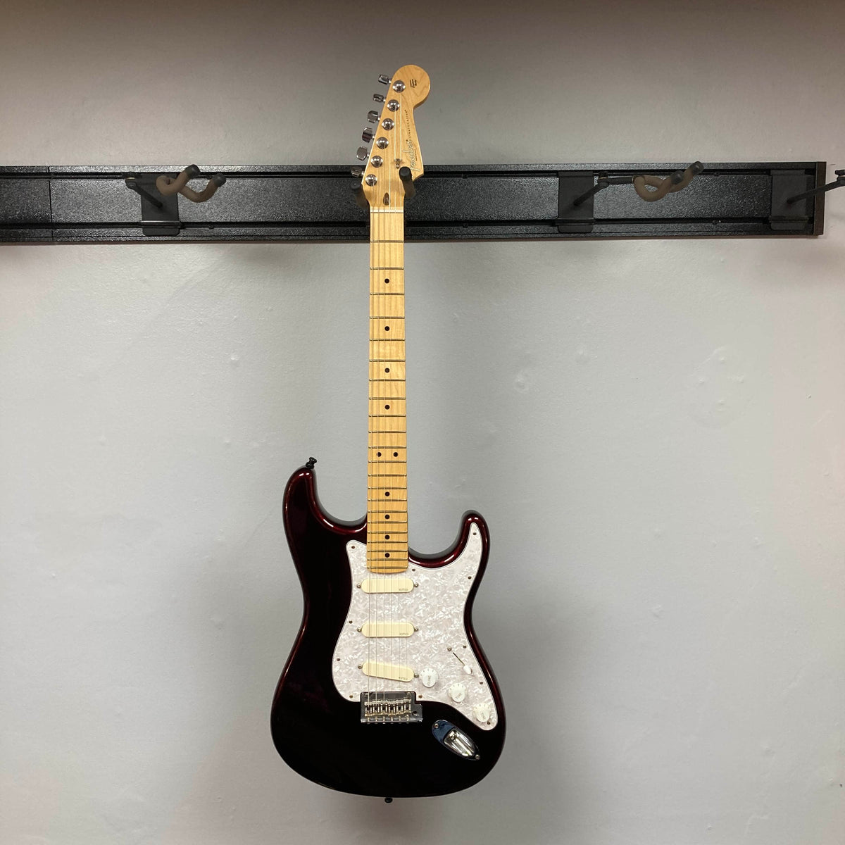 A Fender American Standard Strat upgraded Gilmour EMG DC-20 in striking Bordeaux Metallic, complete with case, is displayed on a wall-mounted rack against a plain light gray wall. The dark-bodied guitar by Fender stands out beautifully with its light pickguard.