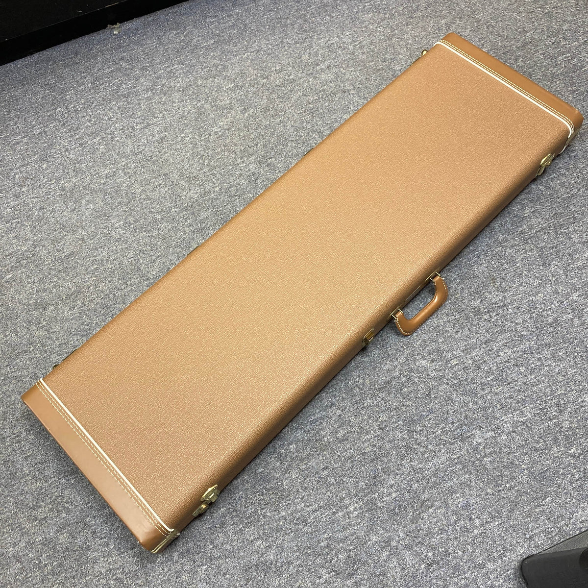 A rectangular brown hard case with handle and metal clasps sits on a gray carpeted floor, designed for the Fender American Vintage '62 Precision Bass Olympic White 2005.