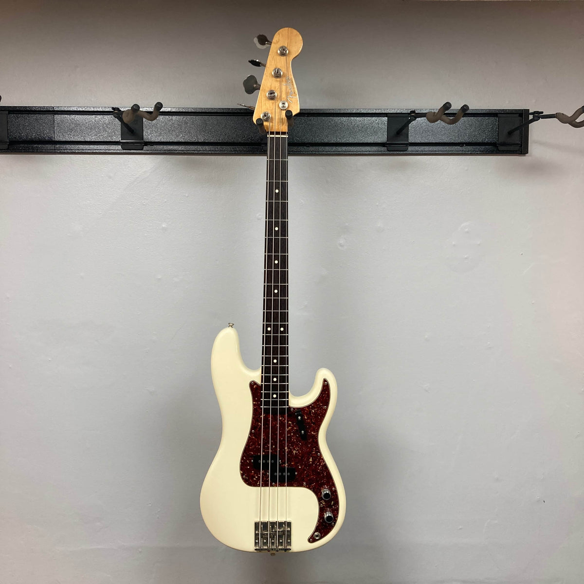 A Fender American Vintage '62 Precision Bass in Olympic White (2005) with a dark red pickguard hangs on a wall-mounted rack against a plain light gray wall.
