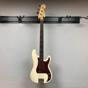 A Fender American Vintage '62 Precision Bass in Olympic White (2005) with a dark red pickguard hangs on a wall-mounted rack against a plain light gray wall.