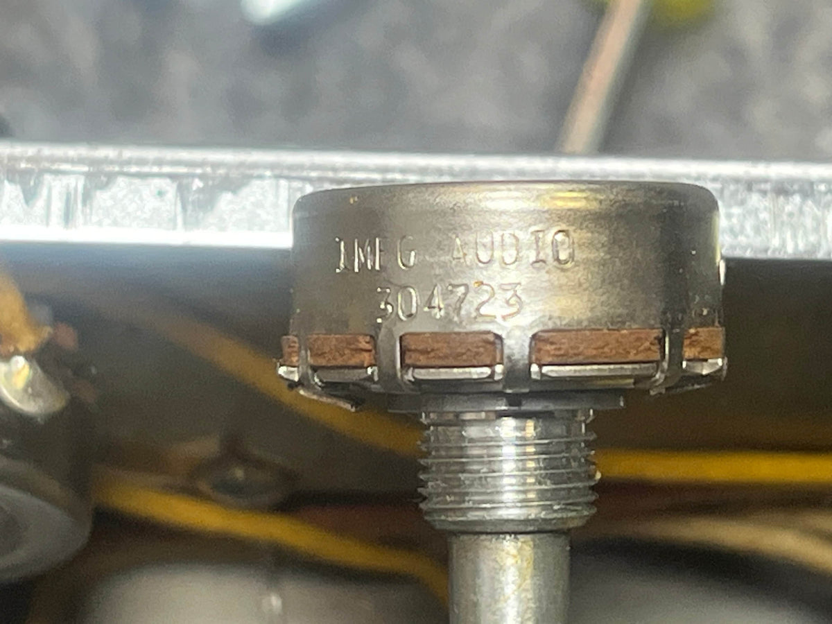 Close-up of a metal component with “MIO A0810 304223” markings and threaded shaft, brown segments, and yellow wires—likely from a Fender 1957 Bassman 5F6 Narrow Panel 40-Watt 4x10" Combo Amp w/Flight Case—in the background.