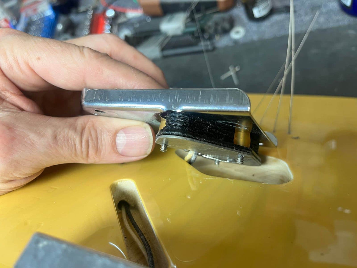 A hand holds a Fender Custom Shop '51 Reissue Nocaster NOS 2006 bridge above a yellow Fender guitar body, with loose wires and components in the background, showcasing vintage Telecaster craftsmanship.