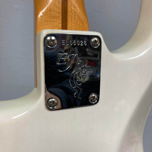 Close-up of a 2006 Fender Eric Johnson Stratocaster neck plate, engraved with "EJ," a serial number, and a flute player figure—secured by four screws to the white body of this signature Fender guitar.