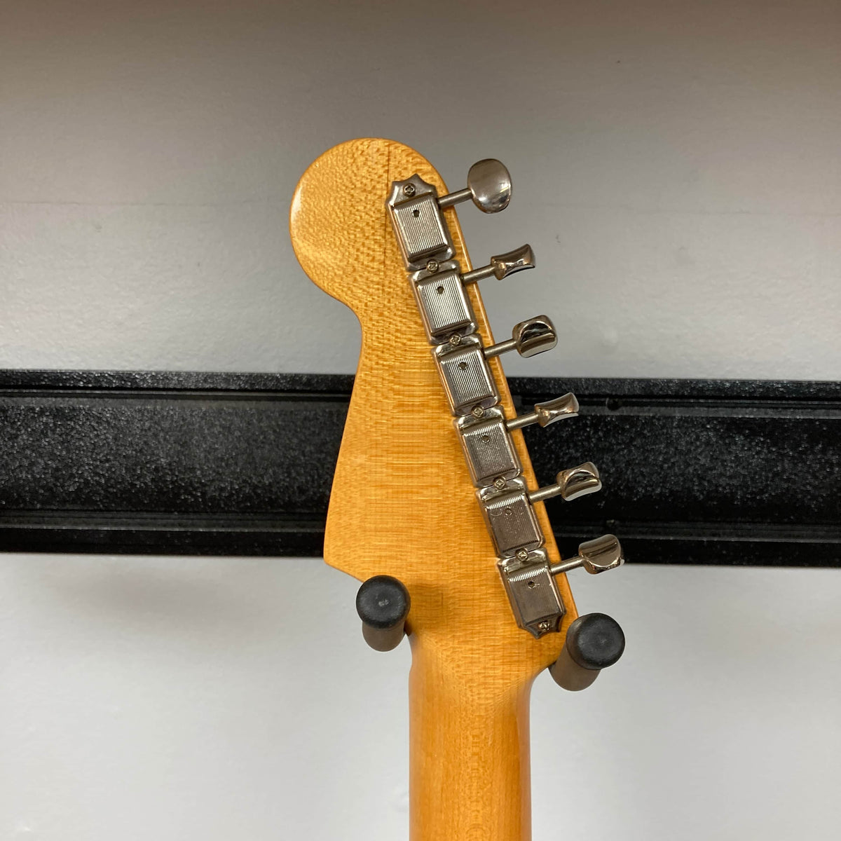 Close-up of the back of a 2006 Fender Eric Johnson Stratocaster headstock by Fender, featuring six metal tuning pegs and two padded hooks securing the maple fingerboard guitar against a wall.
