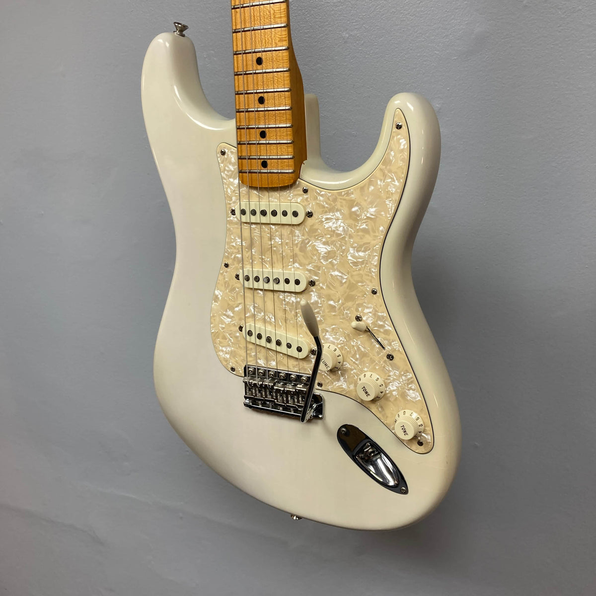 A 2006 Fender Eric Johnson Stratocaster in white, featuring a maple fingerboard, pearloid pickguard, three single-coil pickups, and a tremolo bridge, hangs against a gray wall.