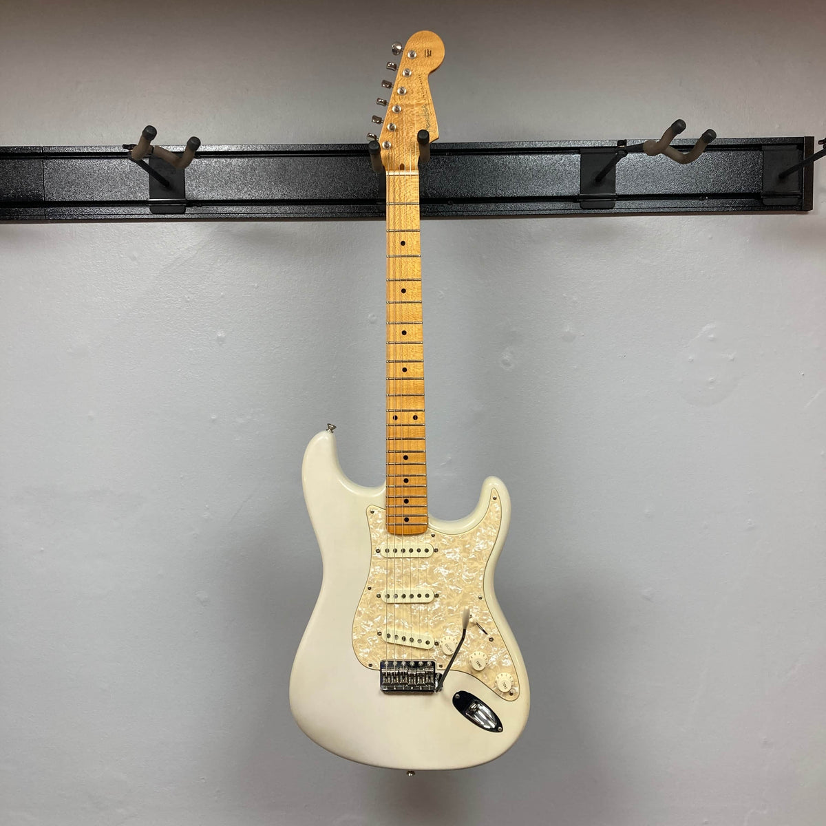 A 2006 Fender Eric Johnson Stratocaster by Fender, featuring a maple fingerboard and neck in white, hangs on a black wall-mounted rack against a light gray wall.