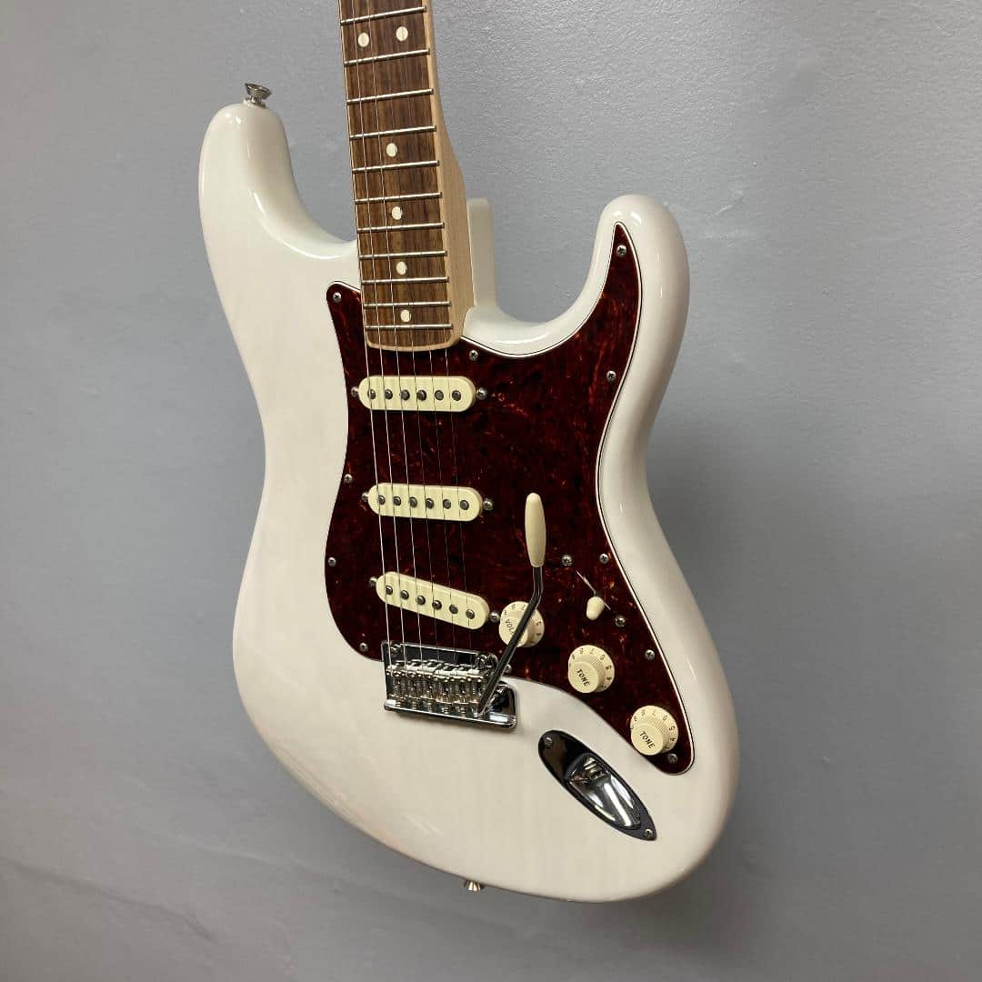 A Fender Limited Edition Ash American Professional Stratocaster in White Blonde with a case hangs on a gray wall, featuring three single-coil pickups and a rosewood fretboard.