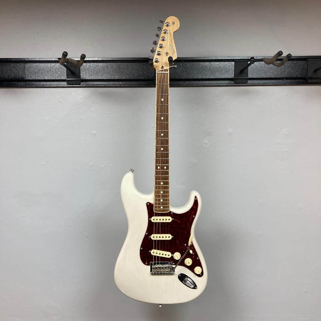 A Fender Limited Edition Ash American Professional Stratocaster in White Blonde with a case hangs against a gray wall.