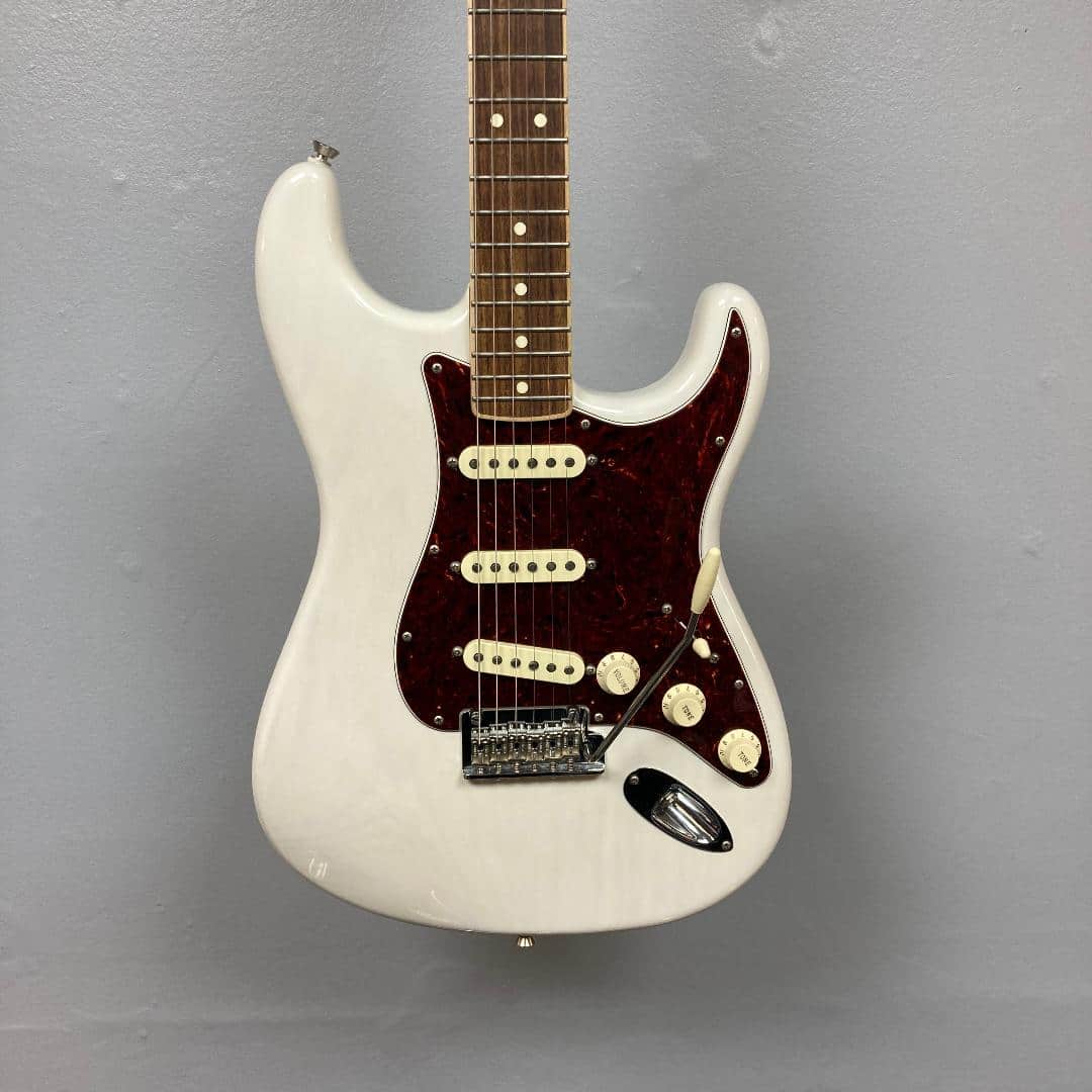 A Fender Limited Edition Ash American Professional Stratocaster in White Blonde with a case hangs against a gray wall.