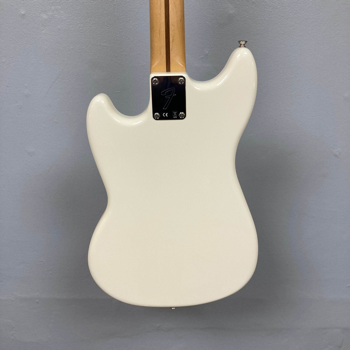 The image shows the back of a Fender Mustang Player Bass PJ White Used, a short scale bass by Fender, with a natural neck secured by a metal plate featuring a stylized &quot;F&quot; and four screws, set against a plain gray wall.