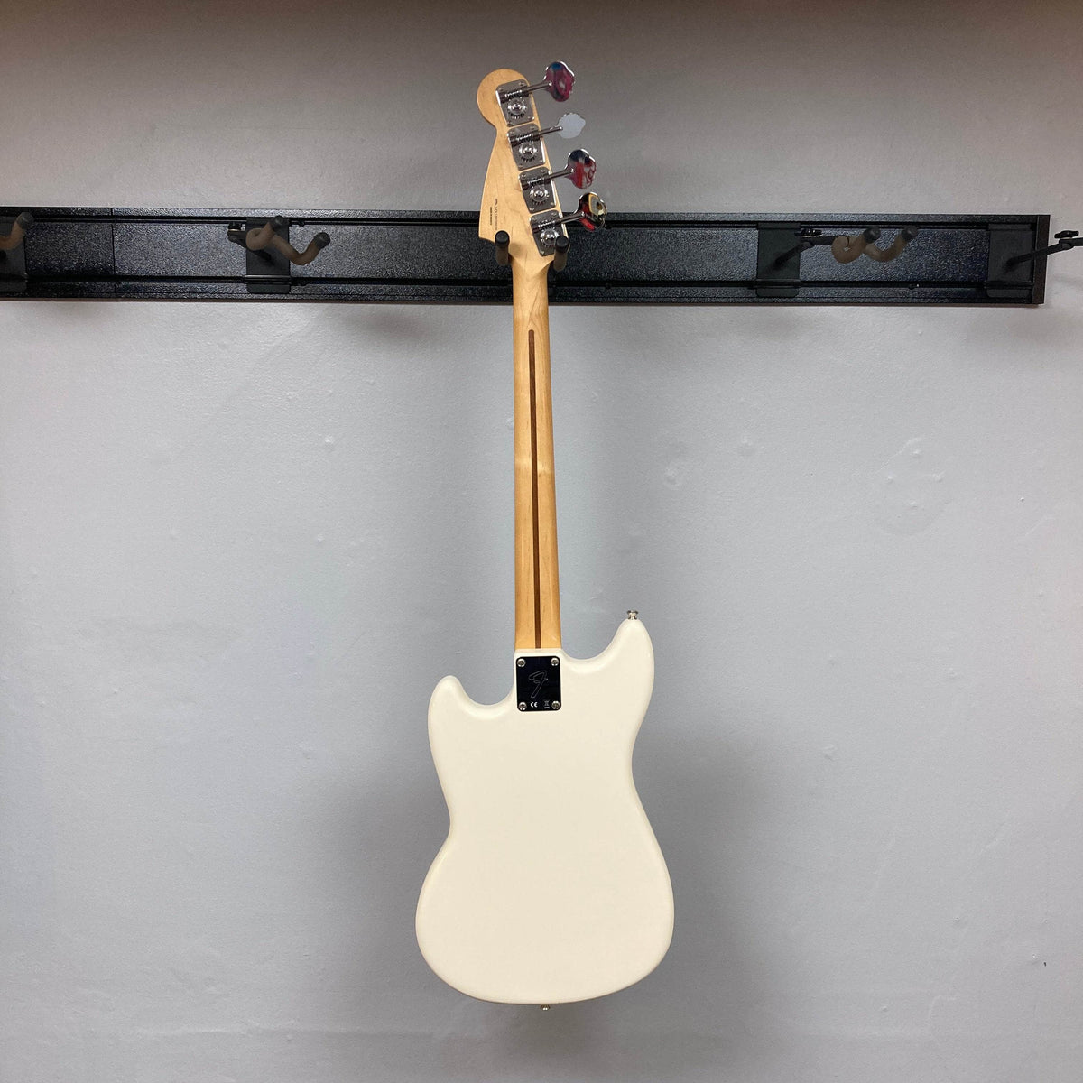 A Fender Mustang Player Bass PJ White Used by Fender hangs on a wall mount with its back, neck, tuning pegs, and backplate visible against a plain, light-colored wall.