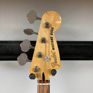 Close-up of the headstock of a used Fender Mustang Player Bass PJ in white, featuring four tuning pegs, the Fender logo, and "Mustang Bass" text, mounted on a wall hanger. Classic short-scale bass by Fender.