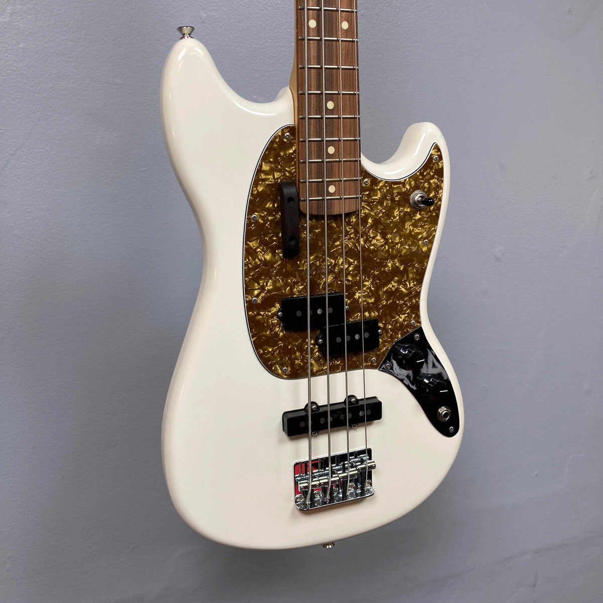 A used Fender Mustang Player Bass PJ in Polar White with a tortoiseshell pickguard, four strings, and chrome hardware from Fender hangs against a gray wall, showcasing its classic short-scale style and compact design.