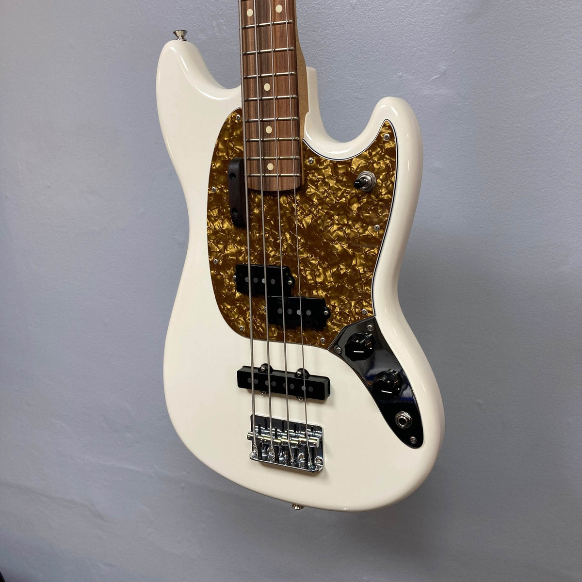 A used Fender Mustang Player Bass PJ in white by Fender, featuring a short scale, brown tortoiseshell pickguard, dual pickups, four knobs, chrome hardware, and displayed against a plain gray wall.