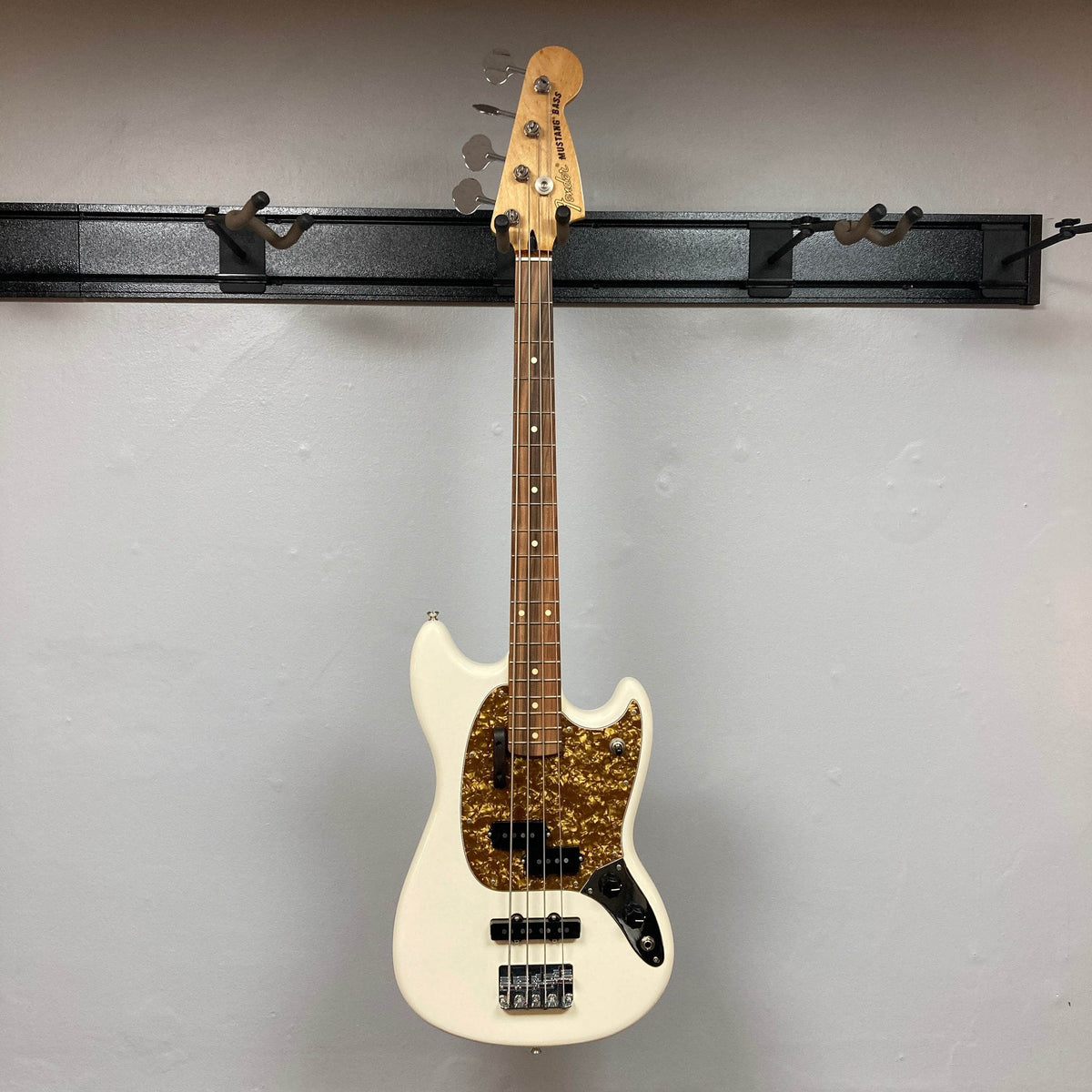 A used Fender Mustang Player Bass PJ in Polar White, featuring a brown tortoiseshell pickguard, hangs on a black wall-mounted rack against a light wall—this stylish short-scale bass by Fender adds character to any space.
