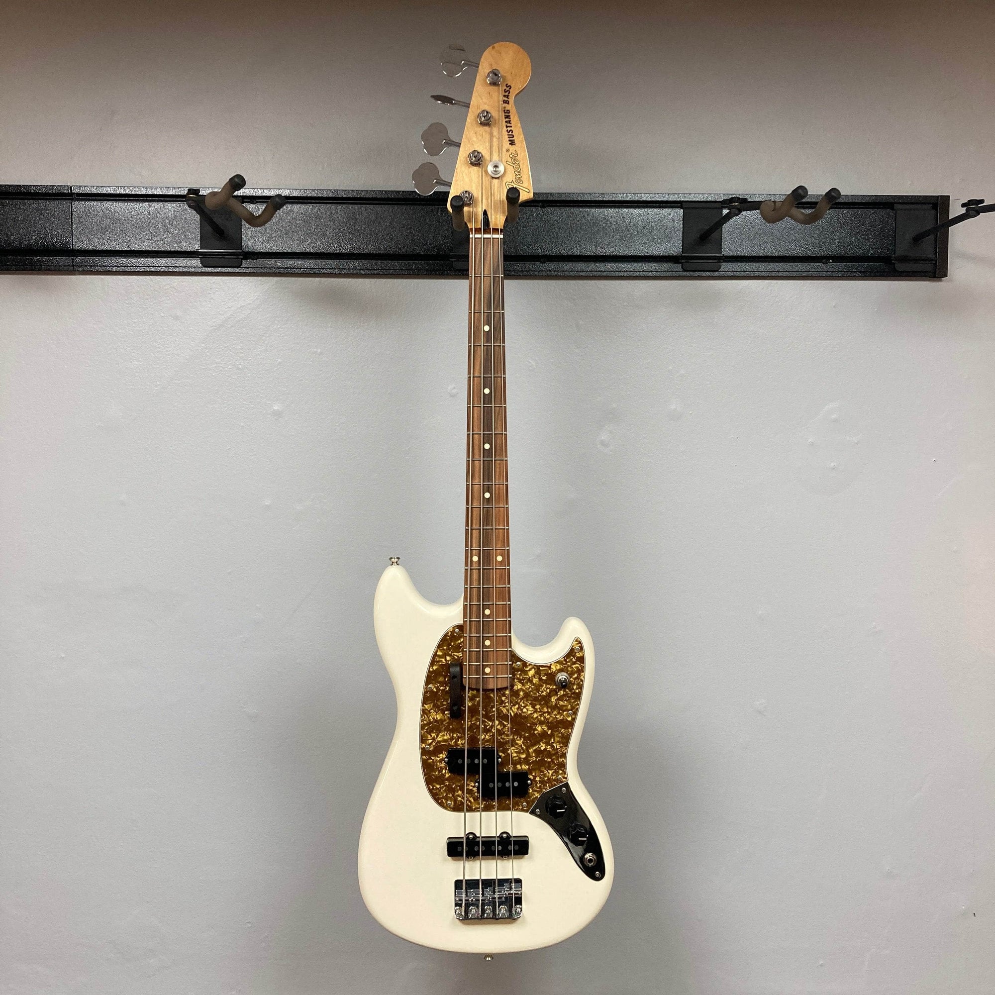 A used Fender Mustang Player Bass PJ in white, this short scale bass by Fender has a brown tortoiseshell pickguard, two pickups, four control knobs, and a wooden fretboard, shown against a plain gray wall.