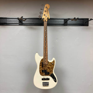 A used Fender Mustang Player Bass PJ in Polar White, featuring a brown tortoiseshell pickguard, hangs on a black wall-mounted rack against a light wall—this stylish short-scale bass by Fender adds character to any space.