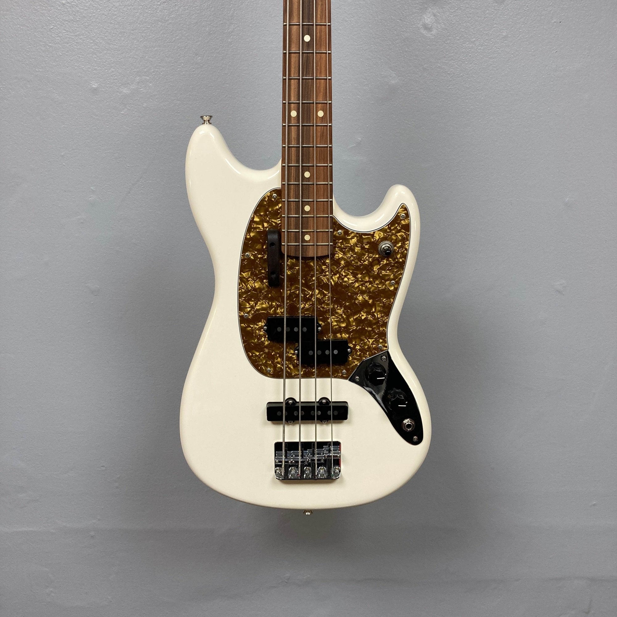 A used Fender Mustang Player Bass PJ in white, this short scale bass by Fender has a brown tortoiseshell pickguard, two pickups, four control knobs, and a wooden fretboard, shown against a plain gray wall.