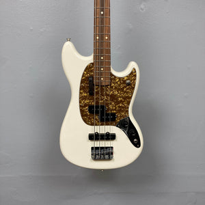 A used Fender Mustang Player Bass PJ in white, this short scale bass by Fender has a brown tortoiseshell pickguard, two pickups, four control knobs, and a wooden fretboard, shown against a plain gray wall.