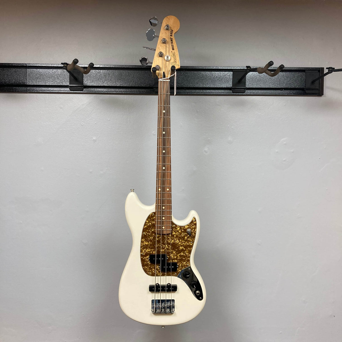 A used Fender Mustang Player Bass PJ in white hangs vertically on a black wall-mounted rack, featuring a brown tortoiseshell pickguard, against a plain light gray wall.