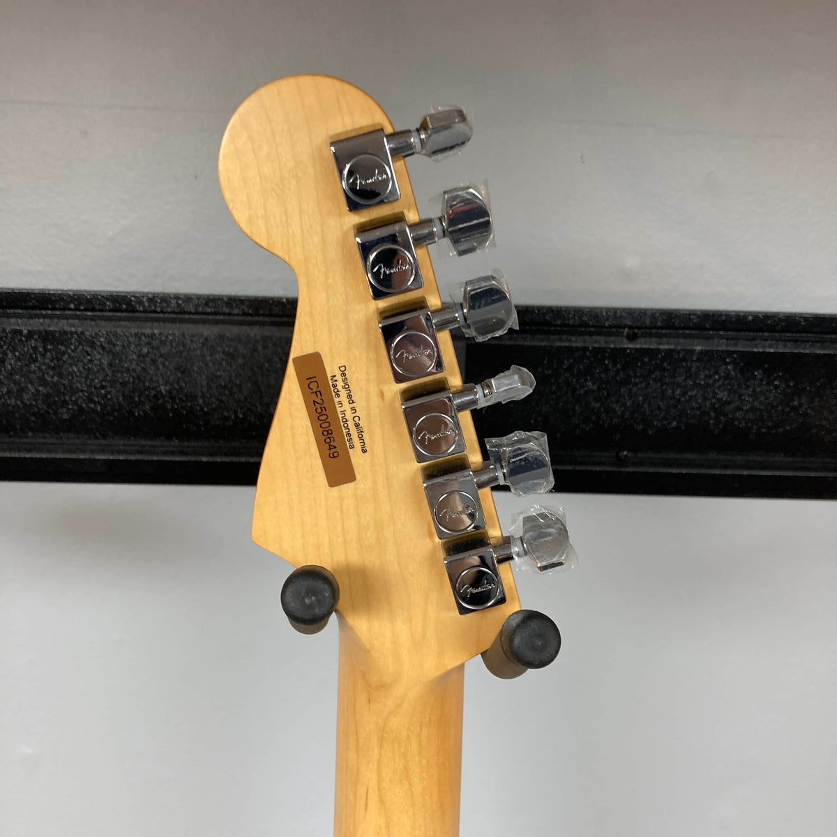 Back of a Fender Standard Stratocaster Olympic White Refurb headstock by Fender, showing six tuning pegs, serial number, and manufacturer info, resting on a wall-mounted guitar holder.