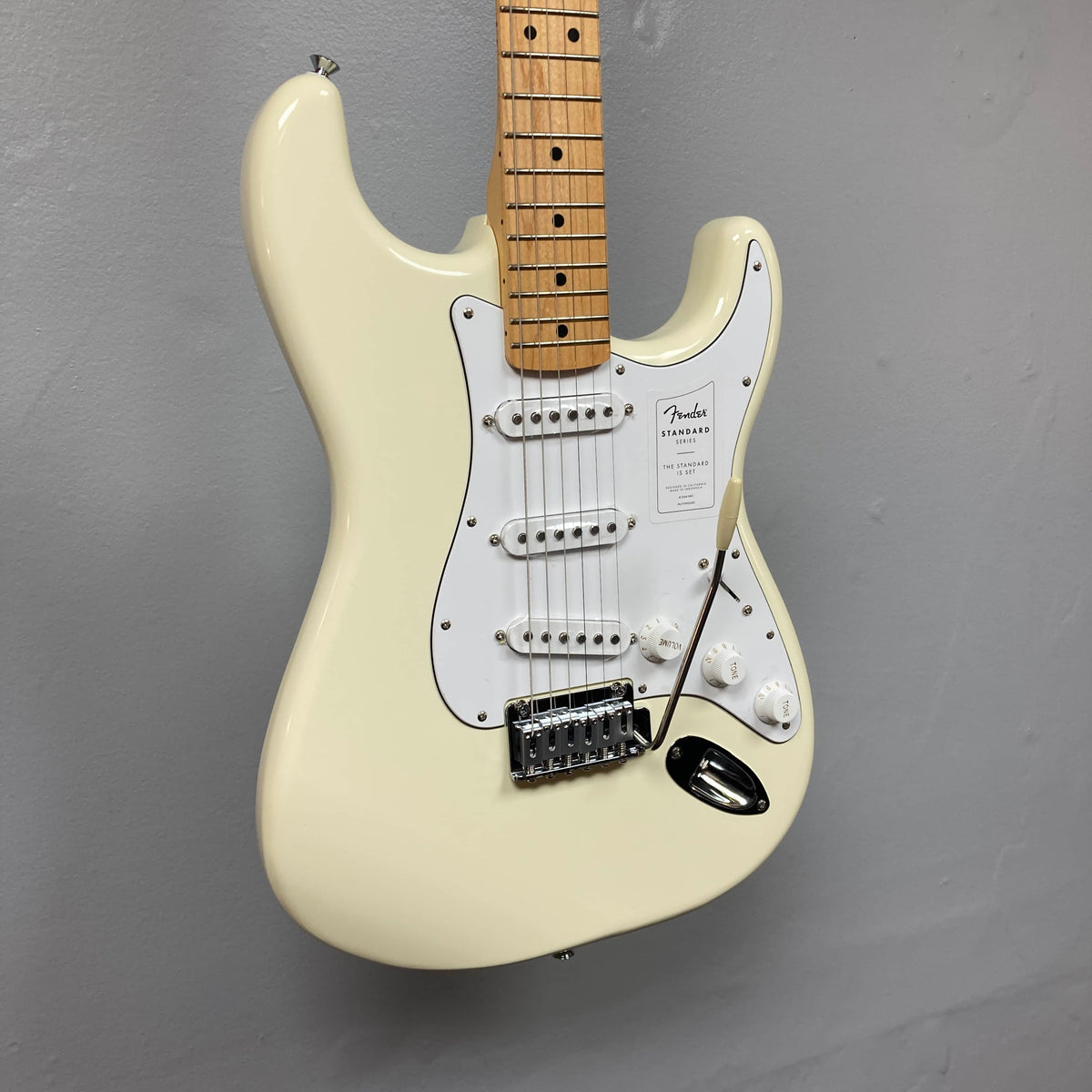 A Fender Standard Stratocaster Olympic White Refurb guitar by Fender, featuring a white finish and maple neck, is mounted against a gray wall.