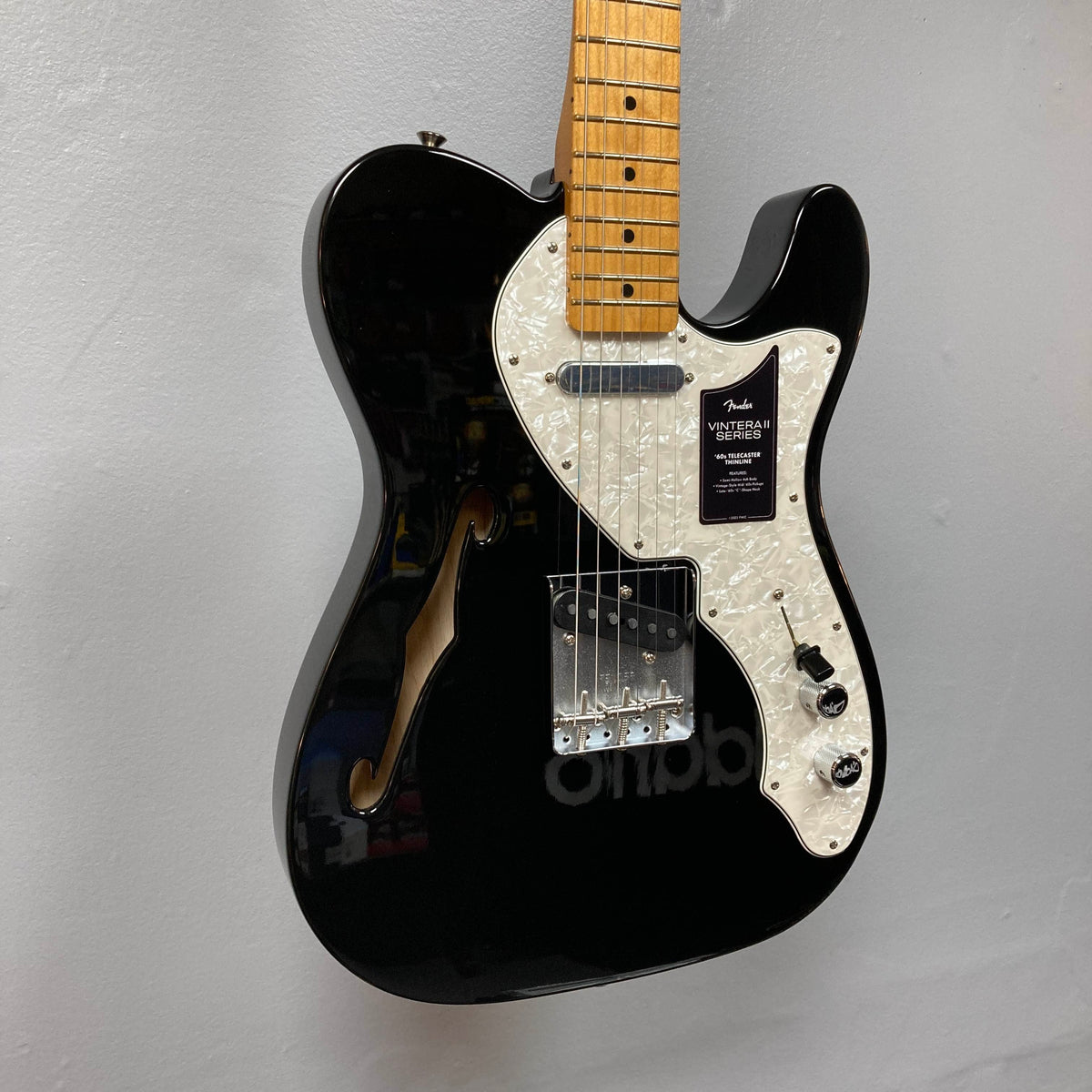 A Fender Vintera II ’60s Telecaster Thinline Black Used electric guitar by Fender, featuring a white pearl pickguard, maple neck, vintage-style Tele pickups, and an f-hole, graces the wall. Its semi-hollow ash body enhances both its classic appearance and resonant tone.
