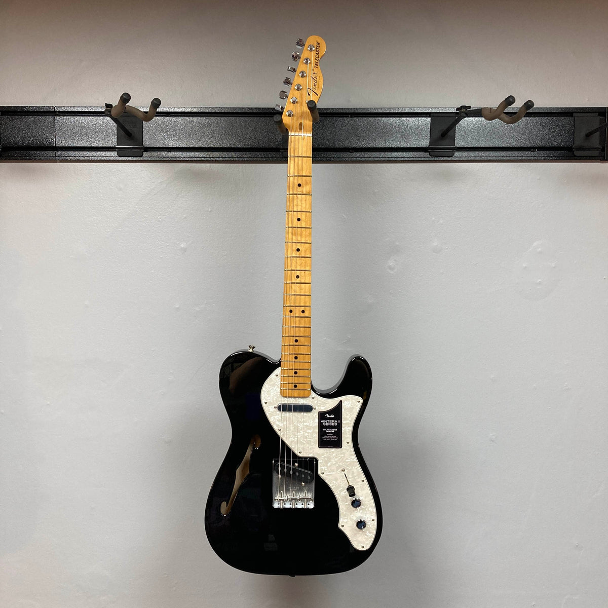 A used Fender Vintera II ’60s Telecaster Thinline electric guitar in black, featuring a semi-hollow ash body, white pickguard, and maple neck, hangs on a wall-mounted rack against a plain white background, displaying its classic vintage-style Tele pickups and iconic Fender design.