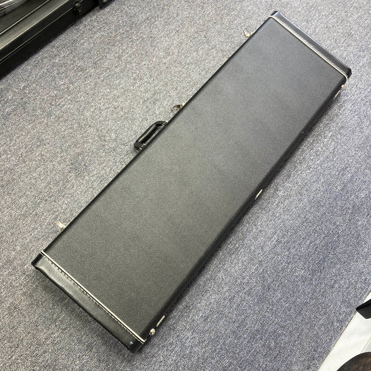 A black hard case with metal latches and a handle sits on a gray carpet, ideal for safely storing your G&L L-2000 USA Tobacco Sunburst Bass (Used Consign).