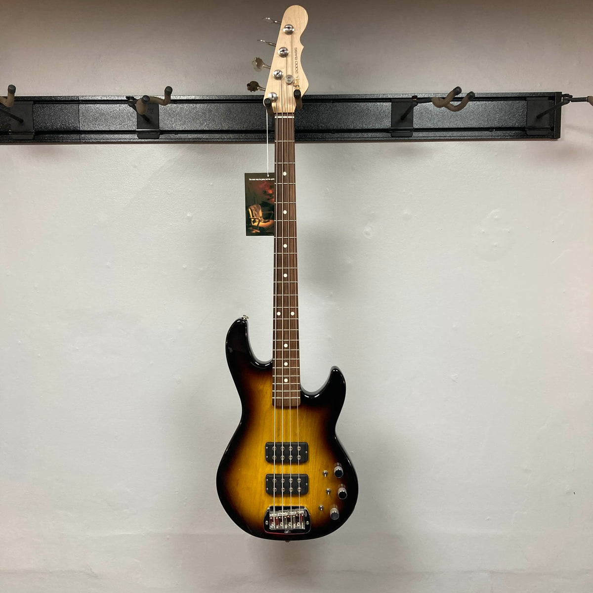 A G&L L-2000 USA Tobacco Sunburst Bass w/Case (Used, Consign) hangs on a black wall-mounted rack against a plain white wall.