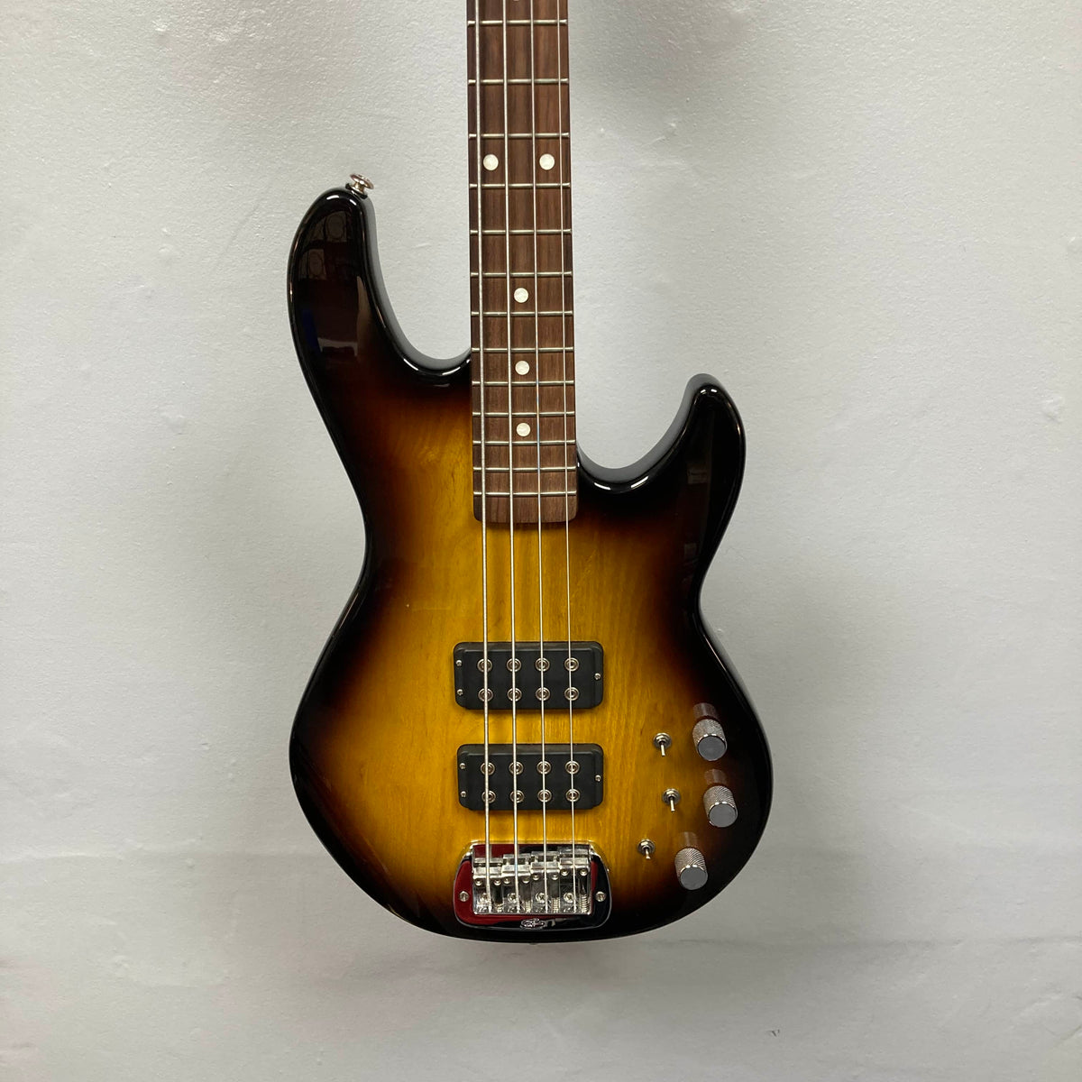 A G&L L-2000 USA Tobacco Sunburst Bass w/Case (used, consign) featuring two pickups and four control knobs rests against a plain, light-colored wall.