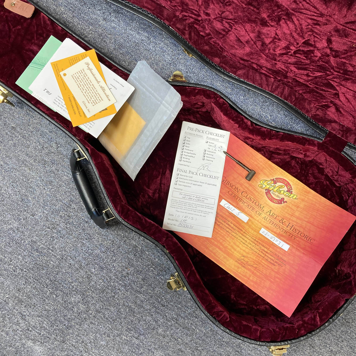 An open guitar case with red velvet lining displays documents including a certificate of authenticity for a Gibson Custom Shop Class 5 Les Paul by Gibson, plus checklists and envelopes, all arranged inside on a gray carpet.