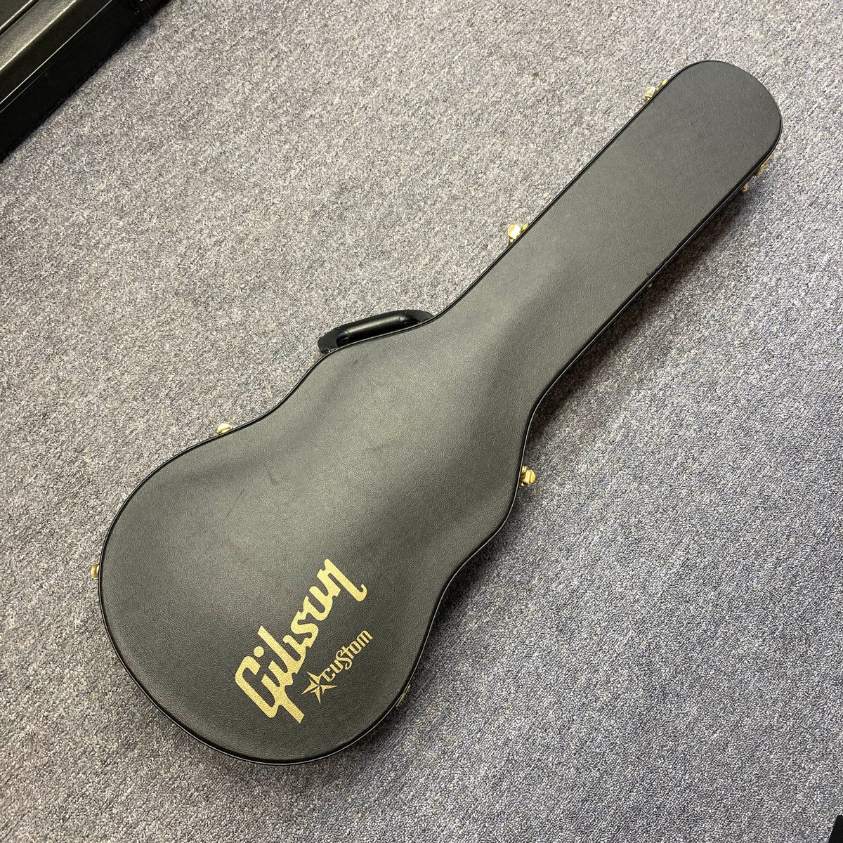 A black Gibson Custom Shop Class 5 Les Paul case with gold lettering rests closed on a gray carpeted floor.