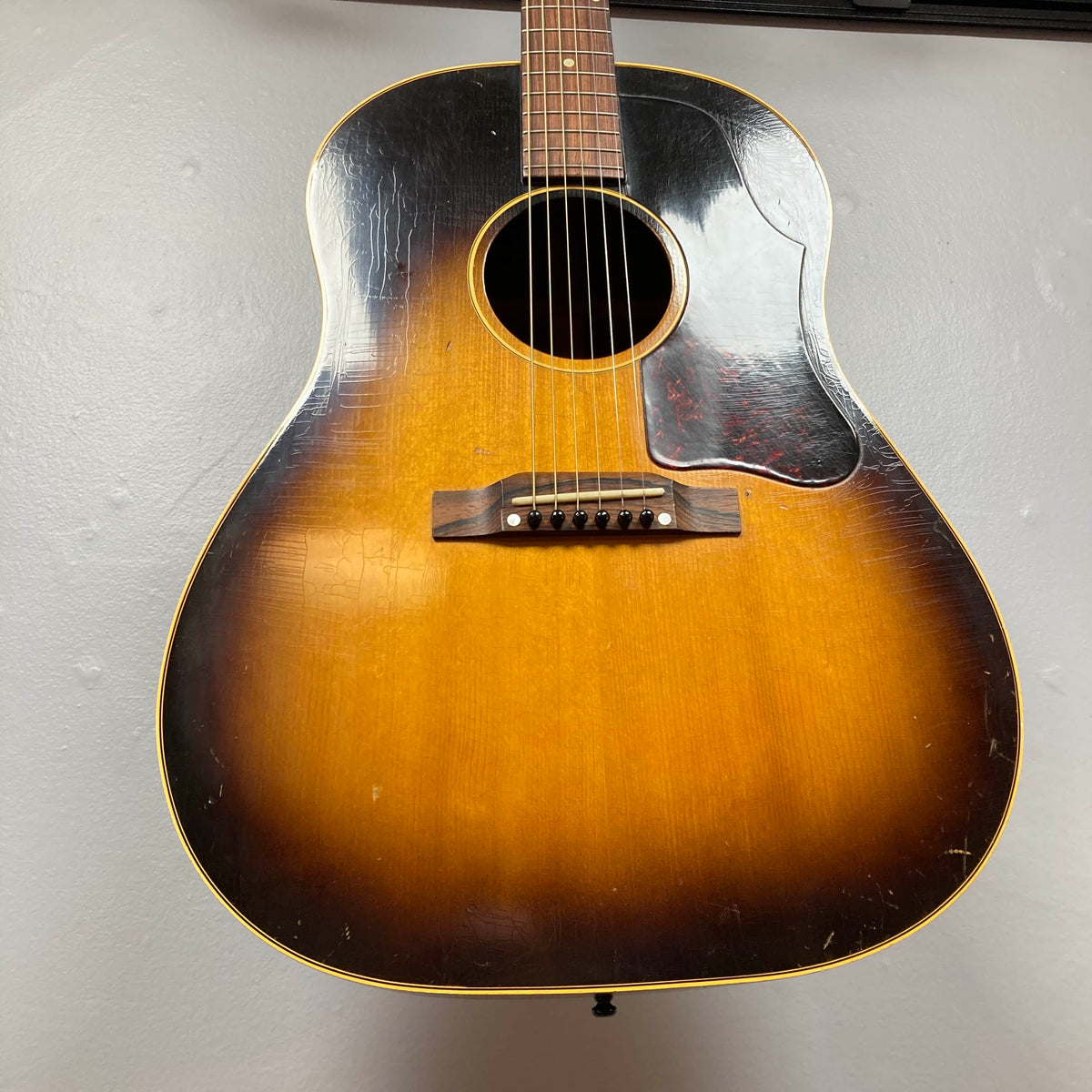 A vintage Gibson J-45 1957 (Serial U-8664-1) with a sunburst finish and worn body hangs on a light-colored wall.