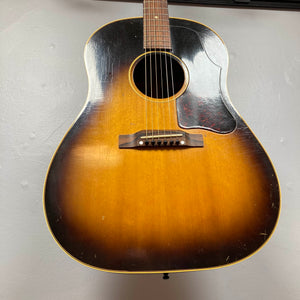 A vintage Gibson J-45 1957 (Serial U-8664-1) with a sunburst finish and worn body hangs on a light-colored wall.
