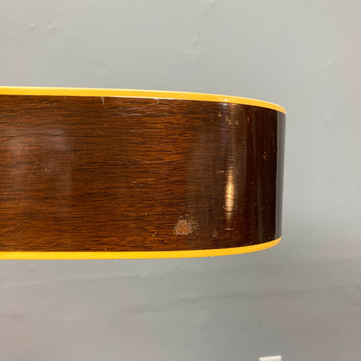 Close-up of the side of a Gibson J-45 1957 (Serial U-8664-1) acoustic guitar, featuring its slope shoulder design, yellow binding, and a small area of worn finish, set against a plain gray wall.