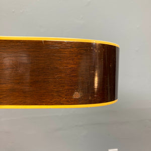 Close-up of the side of a Gibson J-45 1957 (Serial U-8664-1) acoustic guitar, featuring its slope shoulder design, yellow binding, and a small area of worn finish, set against a plain gray wall.