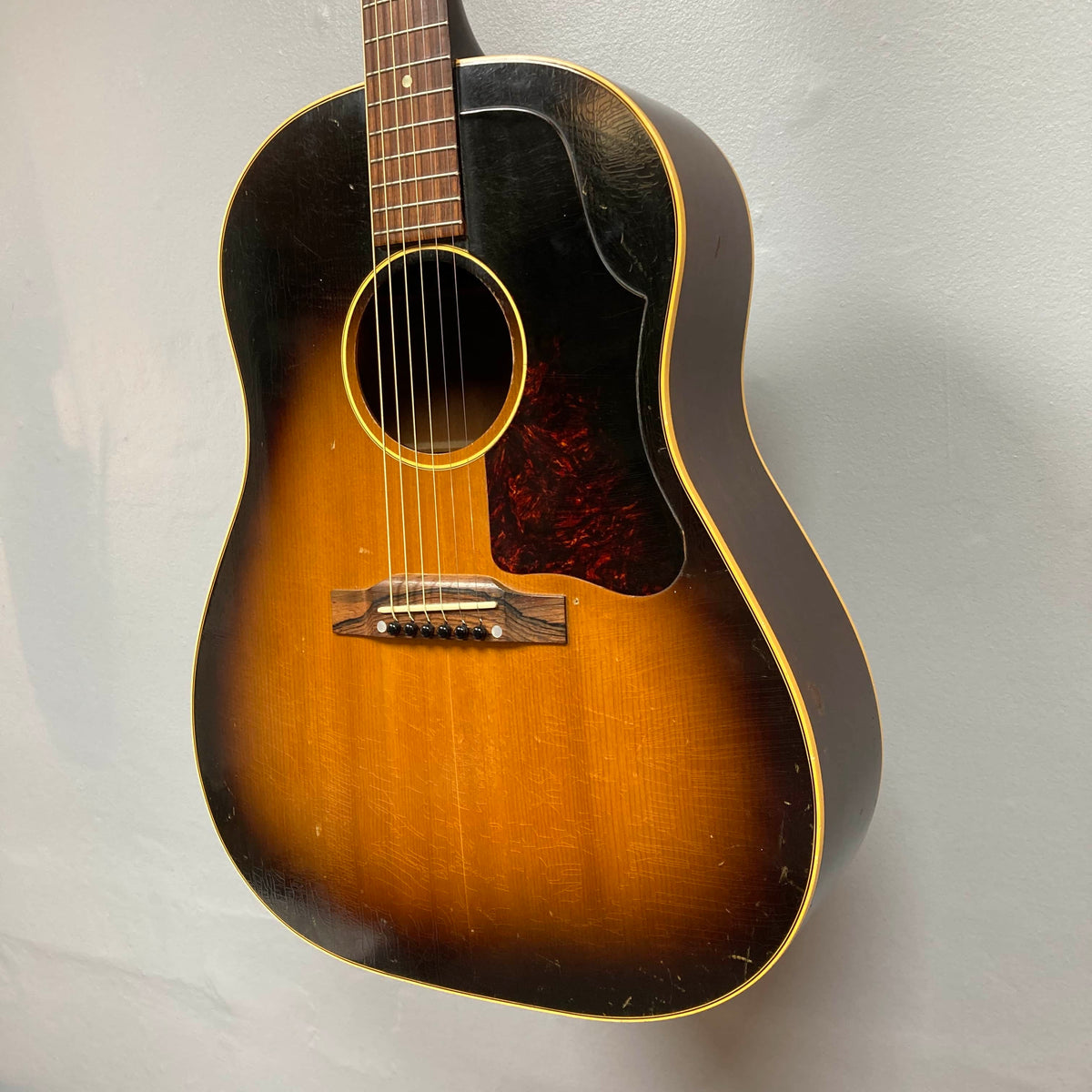 A Gibson J-45 1957 (Serial U-8664-1) vintage acoustic guitar with a sunburst finish and dark pickguard, resembling the classic slope shoulder style, hangs on a white wall.